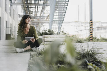 Young smiling woman sitting on the ground and using tablet - KNSF06972