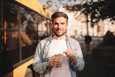 Glücklicher Mann, der eine Tasse Kaffee in der Hand hält, während er im Sommer auf der Straße steht - CAVF71636