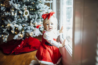 Babymädchen schaut neben Fenster und Weihnachtsbaum in die Kamera - CAVF71710