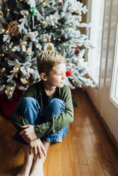 Young boy looking out window next to Christmas tree - CAVF71712