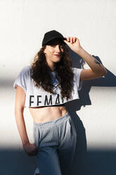 Smiling woman looking at camera leaning on white wall with a cap - CAVF71840