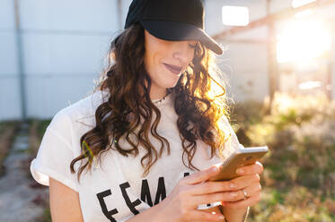 Cheerful millennial woman using smartphone in street at sunset - CAVF71860