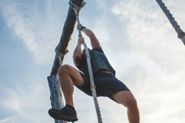 Below-view of a strong male pulling himself up a rope against blue sky - CAVF71987