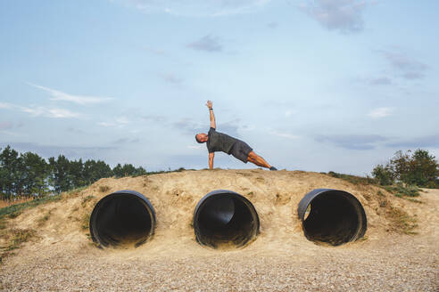 A fit man stretches his arm to the blue sky in a yoga pose on a hill - CAVF71995