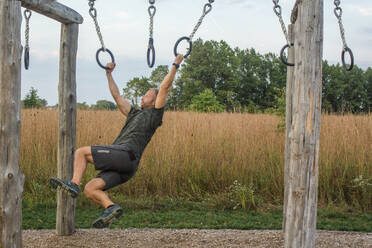 A strong focused man swings from a set of rings outdoors at dusk - CAVF71999