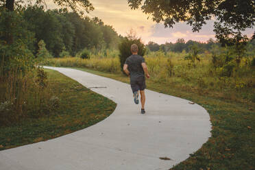 Rear-view of an athletic male running along a path in a park at sunset - CAVF72001