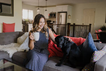 Frau genießt eine Tasse Heißgetränk und streichelt ihren Hund auf dem Sofa - CUF54273