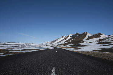 Leere Landstraße an schneebedeckten Bergen vor blauem Himmel - CAVF72432