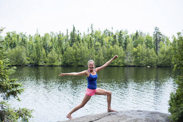 Happy woman practicing yoga while standing on rock at lakeshore against clear sky - CAVF72454