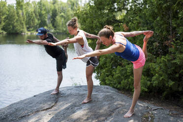 Freunde dehnen ihre Beine beim Yoga auf einem Felsen am Seeufer - CAVF72455