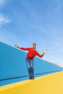 Junger Mann mit Kopfhörern, der Musik hört und auf einer gelben Wand tanzt - AFVF04607