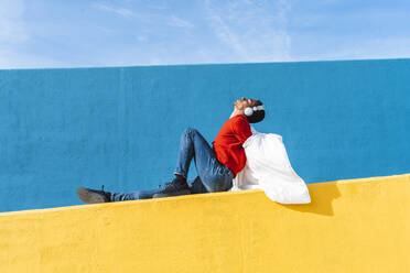 Young dancer sitting on yellow wall, listening music - AFVF04629