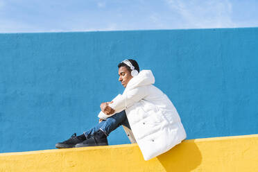 Young trendy man sitting on yellow wall, listening music - AFVF04631