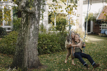 Happy woman embracing senior man on a swing in garden - GUSF02979