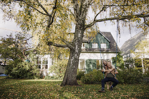 Happy woman embracing senior man on a swing in garden - GUSF02980