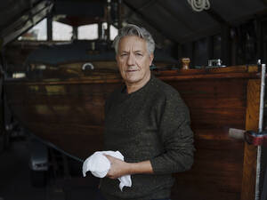 Porträt eines älteren Mannes mit Tuch an einem Holzboot in einem Bootshaus - GUSF02985