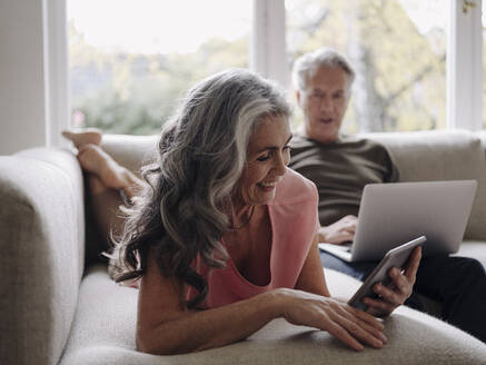 Senior couple relaxing on couch at home using tablet and laptop - GUSF02998