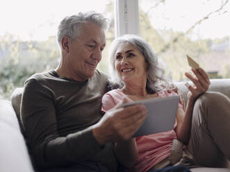 Glückliches älteres Paar, das sich zu Hause auf der Couch entspannt und ein Tablet zum Online-Shopping nutzt - GUSF03001