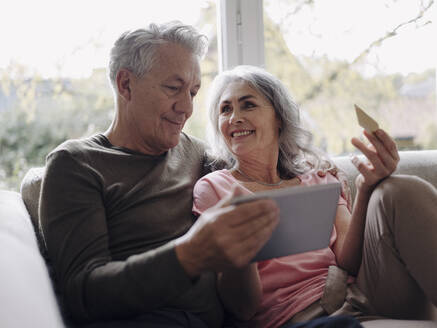 Happy senior couple relaxing on couch at home using tablet for online shopping - GUSF03001