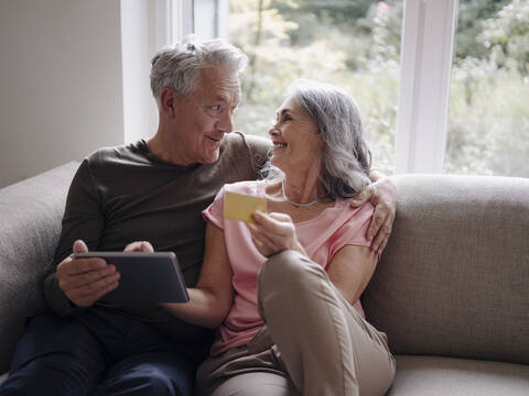 Glückliches älteres Paar, das sich zu Hause auf der Couch entspannt und ein Tablet zum Online-Shopping nutzt, lizenzfreies Stockfoto