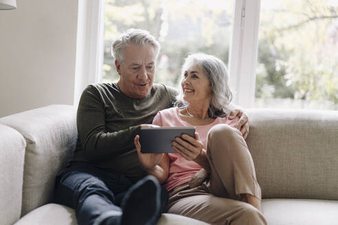 Happy senior couple relaxing on couch at home using tablet - GUSF03056