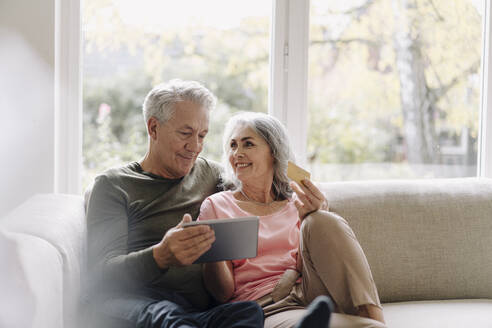 Happy senior couple relaxing on couch at home using tablet for online shopping - GUSF03057