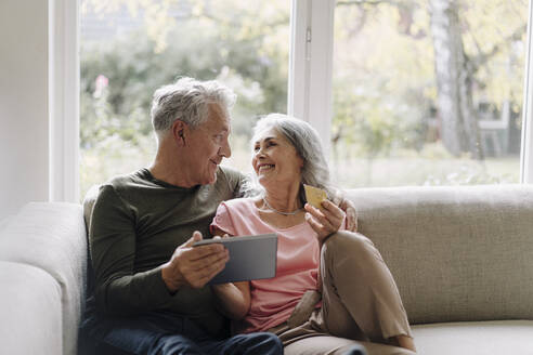 Happy senior couple relaxing on couch at home using tablet for online shopping - GUSF03058