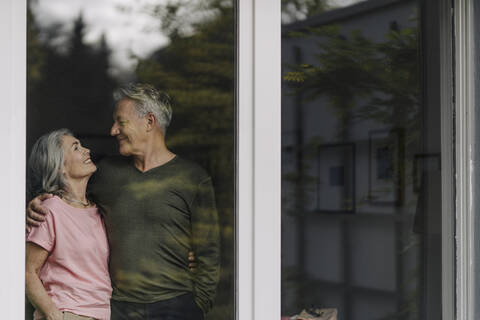 Glückliches Seniorenpaar hinter der Fensterscheibe ihres Hauses, lizenzfreies Stockfoto