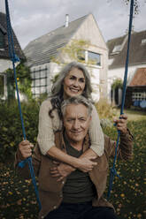 Happy woman embracing senior man on a swing in garden - GUSF03070