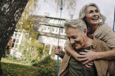 Happy woman embracing senior man on a swing in garden - GUSF03072