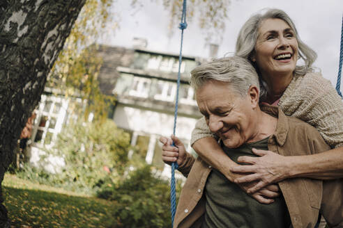 Happy woman embracing senior man on a swing in garden - GUSF03072