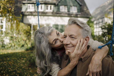 Glückliche Frau umarmt und küsst älteren Mann auf einer Schaukel im Garten - GUSF03073