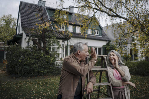 Senior couple with a ladder high fiving in garden of their home - GUSF03077