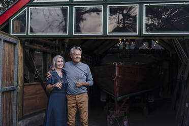 Senior couple standing in front of boathouse with glass of champagne - GUSF03087