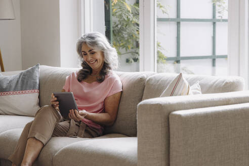 Lächelnde reife Frau mit Tablet auf der Couch sitzend zu Hause - GUSF03106