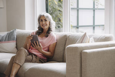 Porträt einer lächelnden reifen Frau mit Tablet auf der Couch zu Hause sitzen - GUSF03107