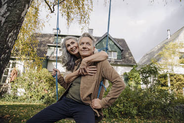 Happy woman embracing senior man on a swing in garden - GUSF03143