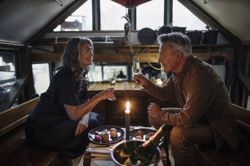 Senior couple having a candlelight dinner on a boat in boathouse clinking champagne glasses - GUSF03153