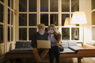 Senior couple using laptop on couch at home at night - GUSF03166