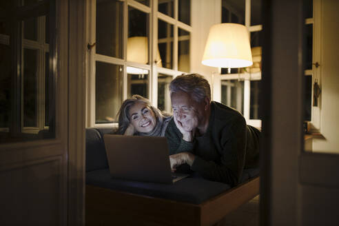 Glückliches älteres Paar, das nachts zu Hause auf der Couch liegt und einen Laptop benutzt - GUSF03171