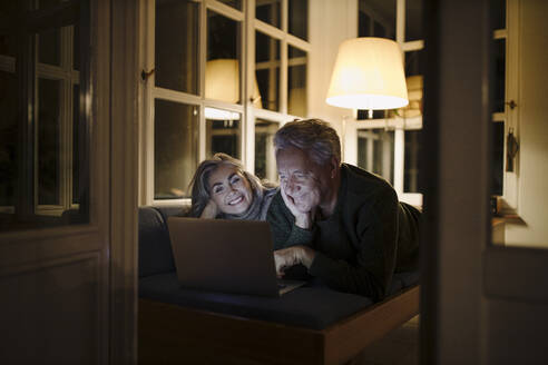 Happy senior couple lying on couch at home at night using laptop - GUSF03171