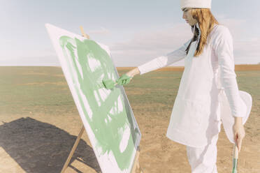 Young woman on dry field, painting canvas with green paint - ERRF02344