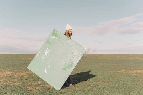 Young woman holding green painting on dry field - ERRF02383