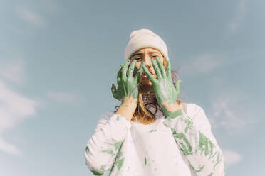 Young woman holding green painted hands in front of her face - ERRF02387