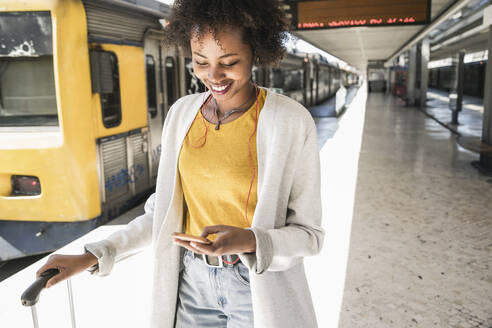 Lächelnde junge Frau mit Kopfhörern und Smartphone am Bahnsteig - UUF19741