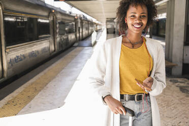 Porträt einer lächelnden jungen Frau mit Kopfhörern und Smartphone am Bahnsteig - UUF19742