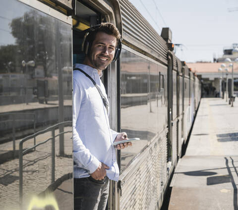 Lächelnder junger Mann mit Kopfhörern und Smartphone in der Zugtür stehend, lizenzfreies Stockfoto