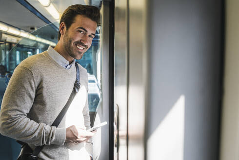 Porträt eines lächelnden jungen Mannes mit Smartphone in einem Zug - UUF19777