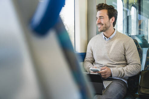 Smiling young man with smartphone and tablet on a train - UUF19781