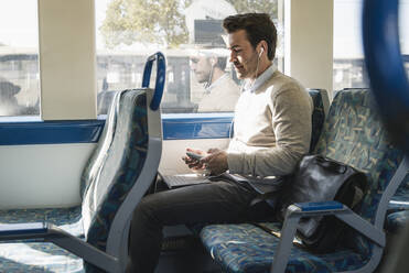 Young man with earphones using smartphone and tablet on a train - UUF19784